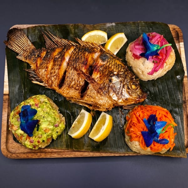 Whole fried fish with lemon, guacamole, pickled onions, and shredded carrots on banana leaves.